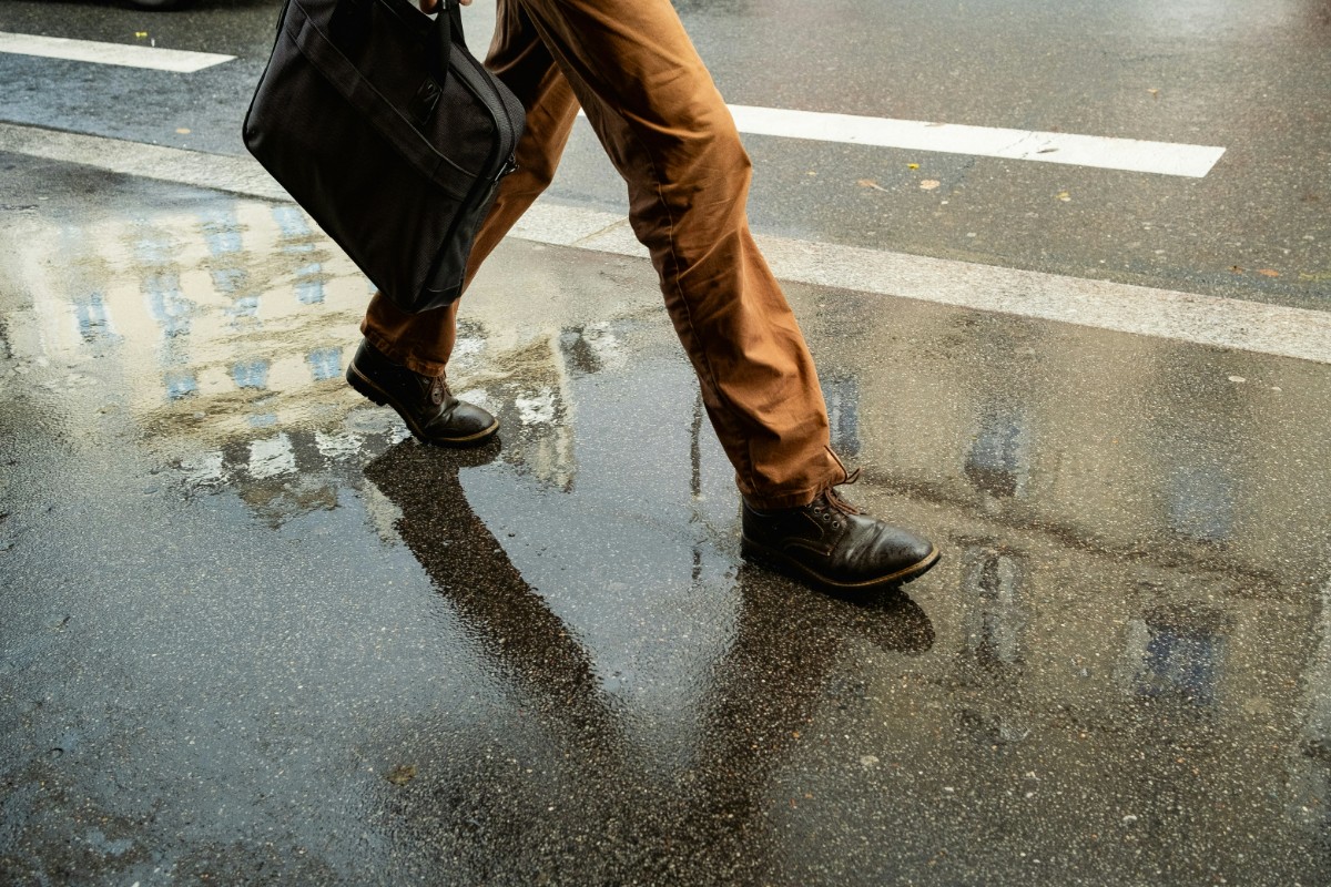 Person in brown pants and black shoes walking on wet pavement, carrying a black bag. Reflections of nearby buildings visible in the puddles.