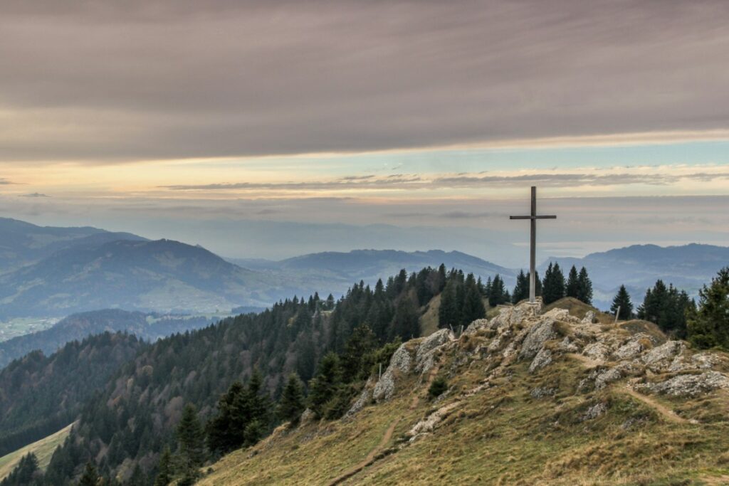A cross stands on a rocky hilltop, surrounded by pine trees, overlooking a panoramic view of distant mountains under a cloudy sky.