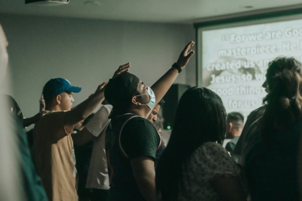 People in a room, facing a screen with text, some holding their hands up.