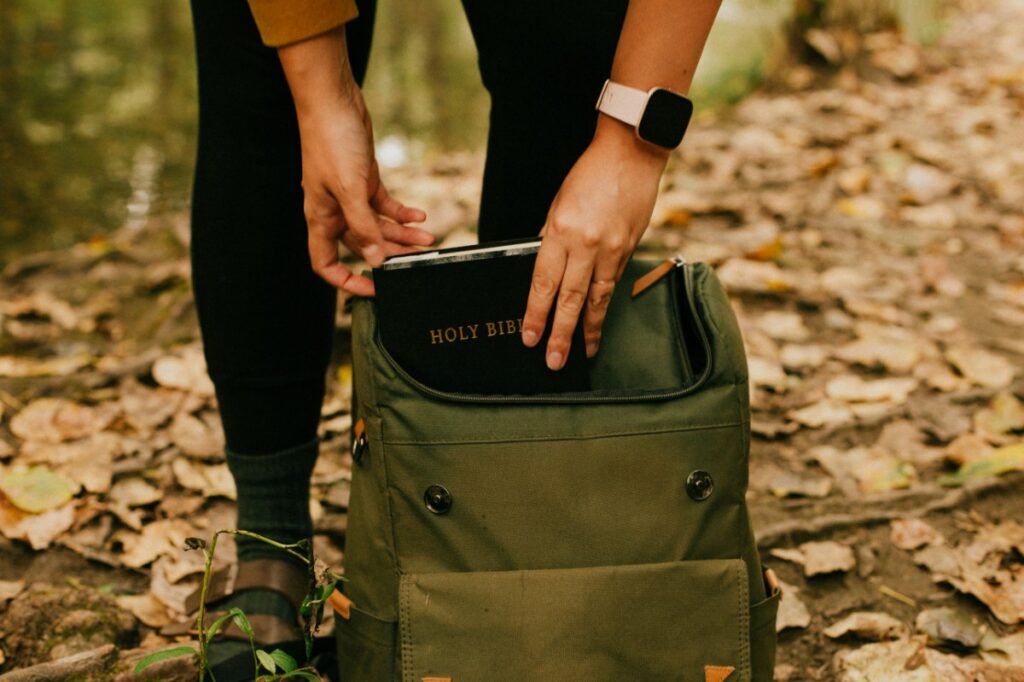 Person placing a book titled Holy Bible into an olive green backpack outdoors, surrounded by fallen leaves.