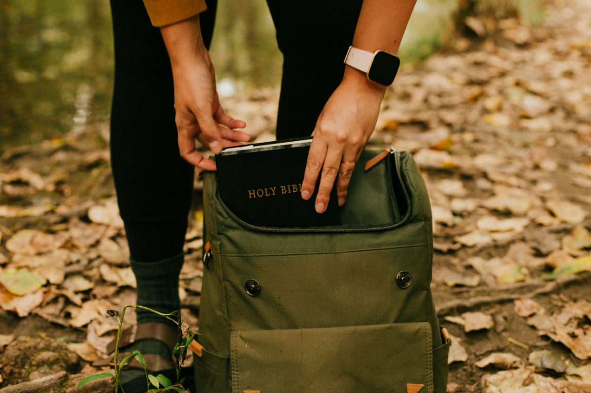 Person placing a book titled Holy Bible into an olive green backpack outdoors, surrounded by fallen leaves.