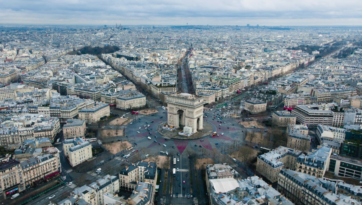 Aerial view of the Arc de Triomphe in Paris, with streets radiating outwards, surrounded by city buildings under a cloudy sky.