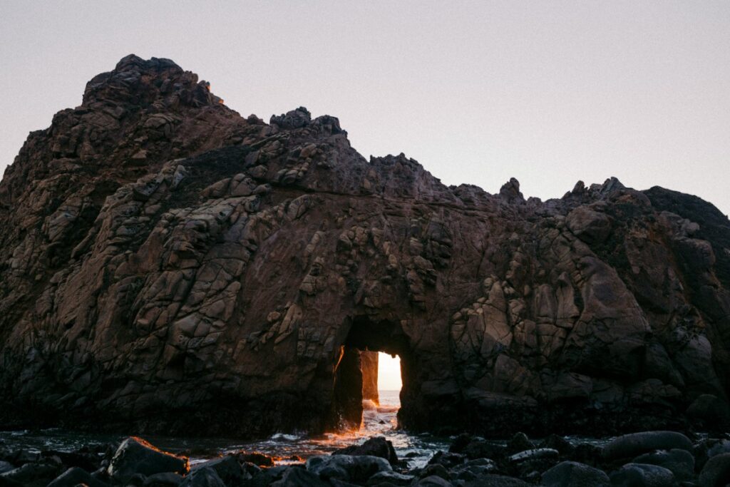 A rocky formation with an arched opening, allowing sunlight to shine through, surrounded by dark water and a clear sky.