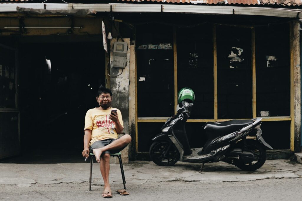 A man sits on a chair in front of a dark building, looking at his phone. A parked scooter is next to him.