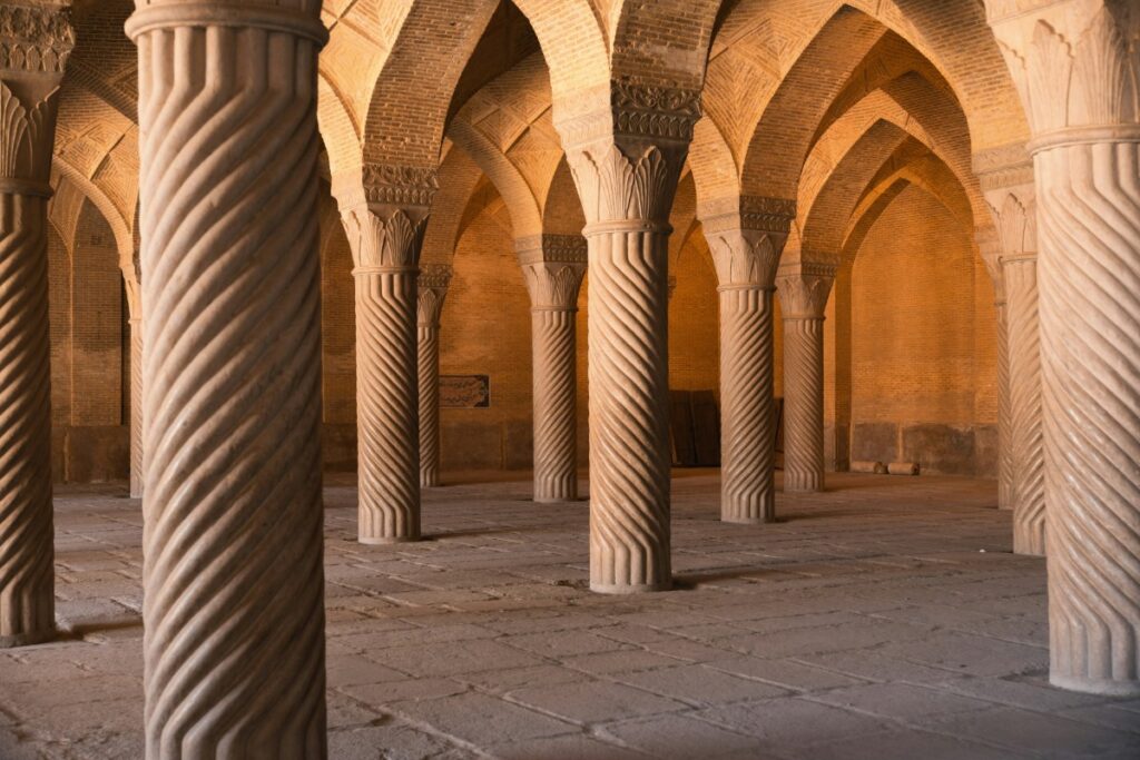 Interior of an ancient structure with intricately carved, spiraled stone columns and arches, lit by warm sunlight.