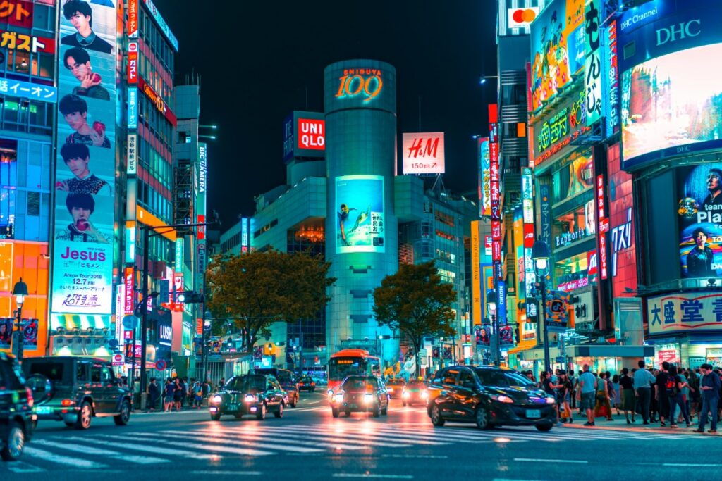 Busy city street at night with illuminated billboards and neon signs. Cars and people fill the scene, creating a vibrant and lively urban atmosphere.
