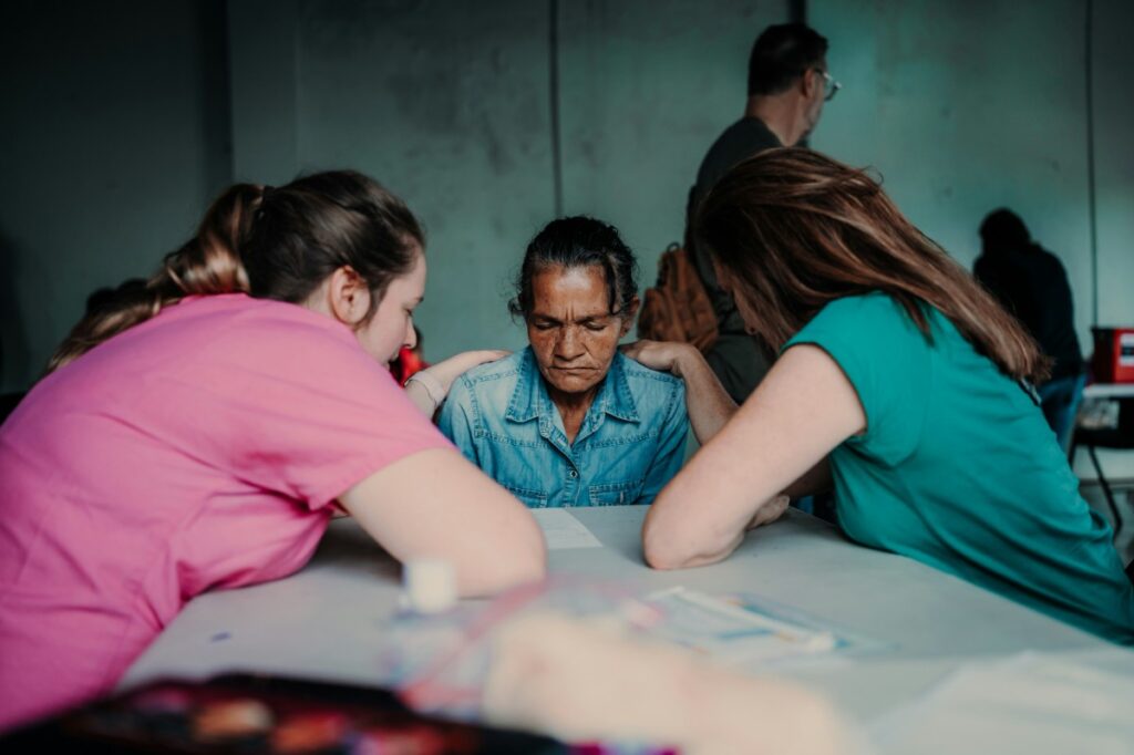 Three people sitting at a table, two women facing a man in the center. The room is dimly lit.