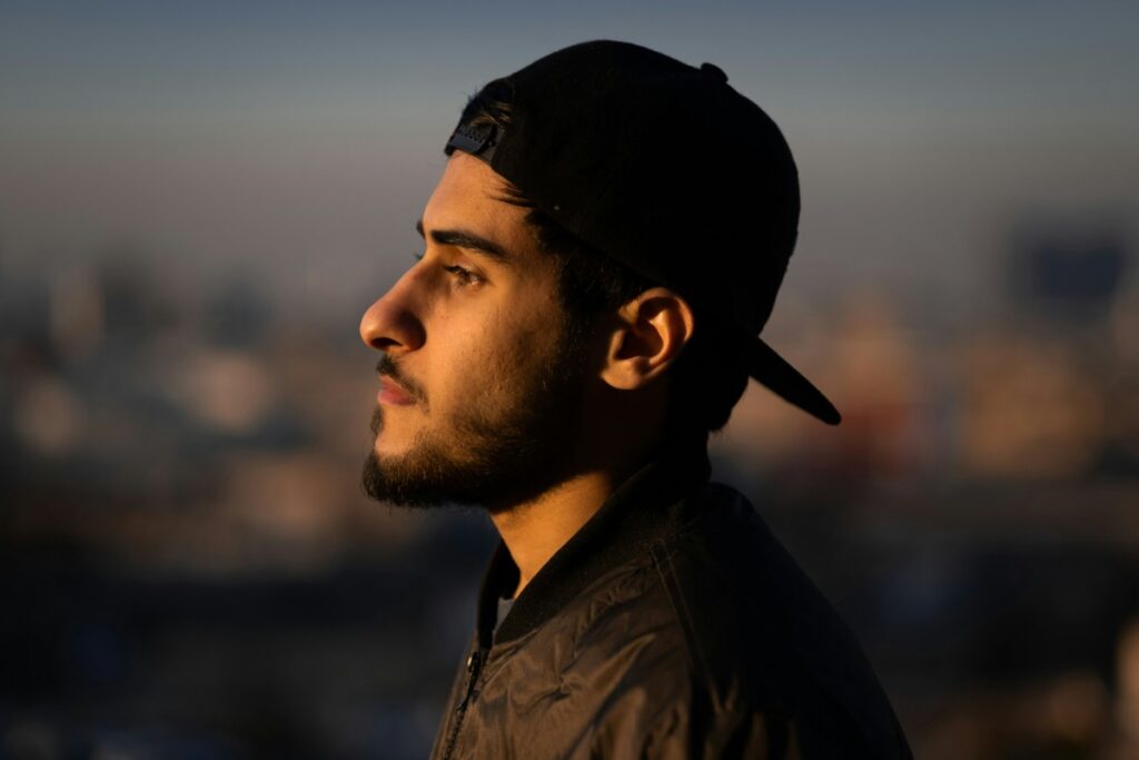 Side profile of a person with a beard wearing a cap and jacket, looking into the distance against a blurred urban background.