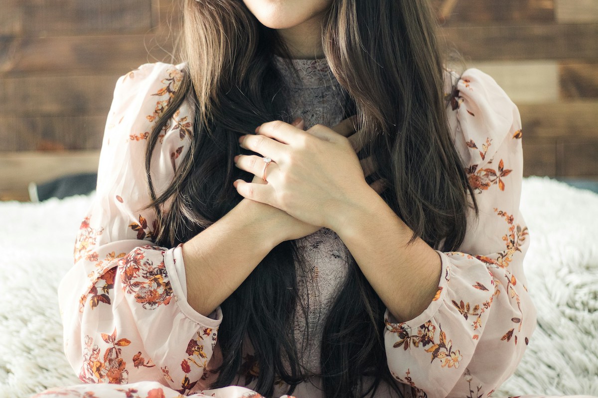 A female with long dark hair, wearing a floral dress, sits serenely against a wooden wall background, her hands clasped over her chest in a gesture that evokes spirituality.
