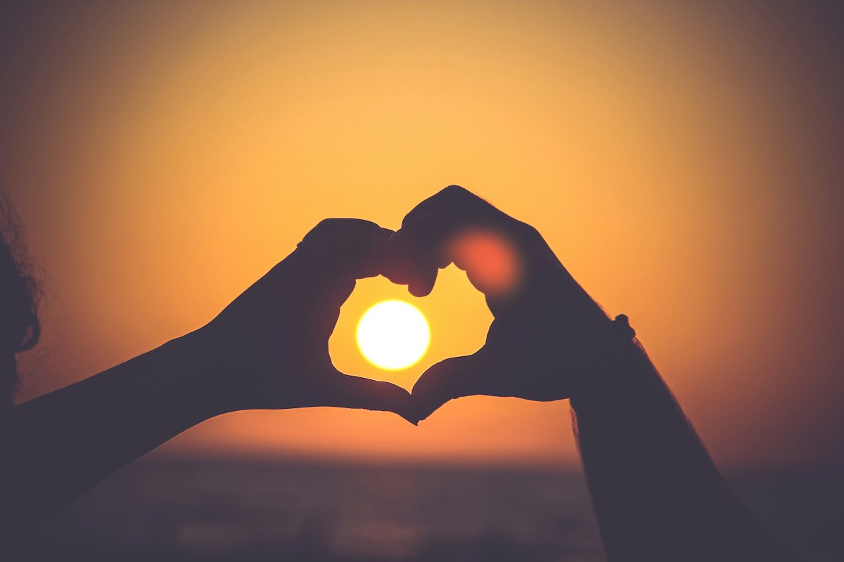 Two hands form a heart shape against a sunset background, capturing the essence of love as they frame the sun at the center.