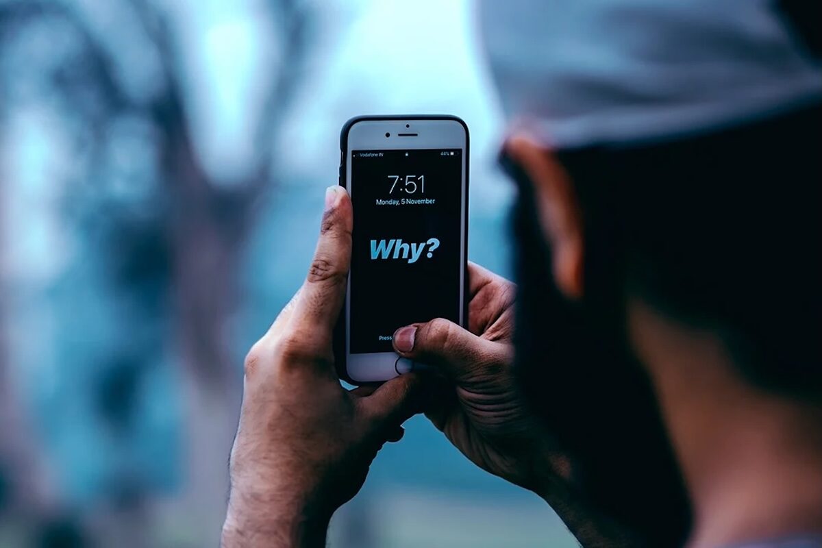 Person holding a smartphone displaying the time 7:51 and the word Why? on the screen, with a blurred outdoor background.