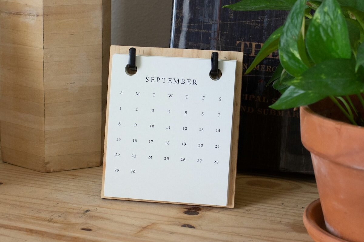 A small desk calendar open to September is placed on a wooden surface next to a potted plant and books.