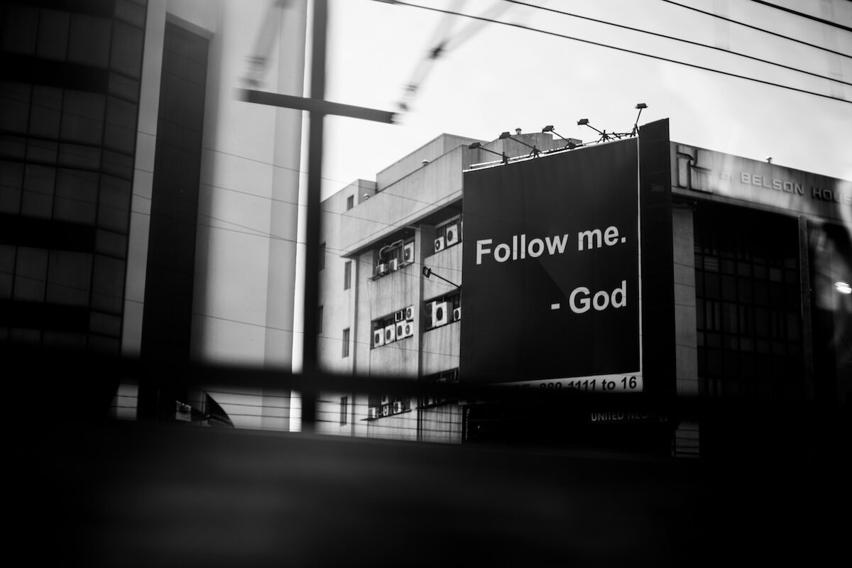 A black and white photo shows a large billboard on a city building with the text, Follow me. - God. Power lines cross in the foreground.