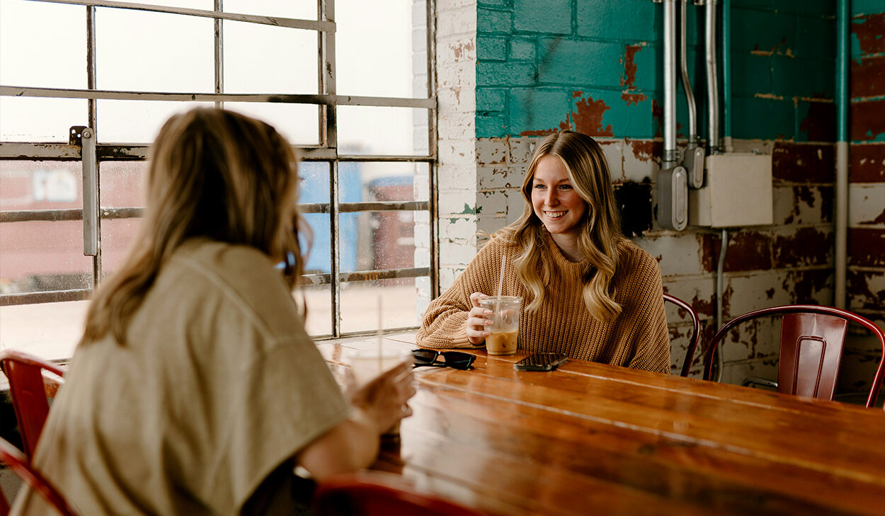 Two blonde women are sitting at a table at a coffee shop, talking and drinking coffee.