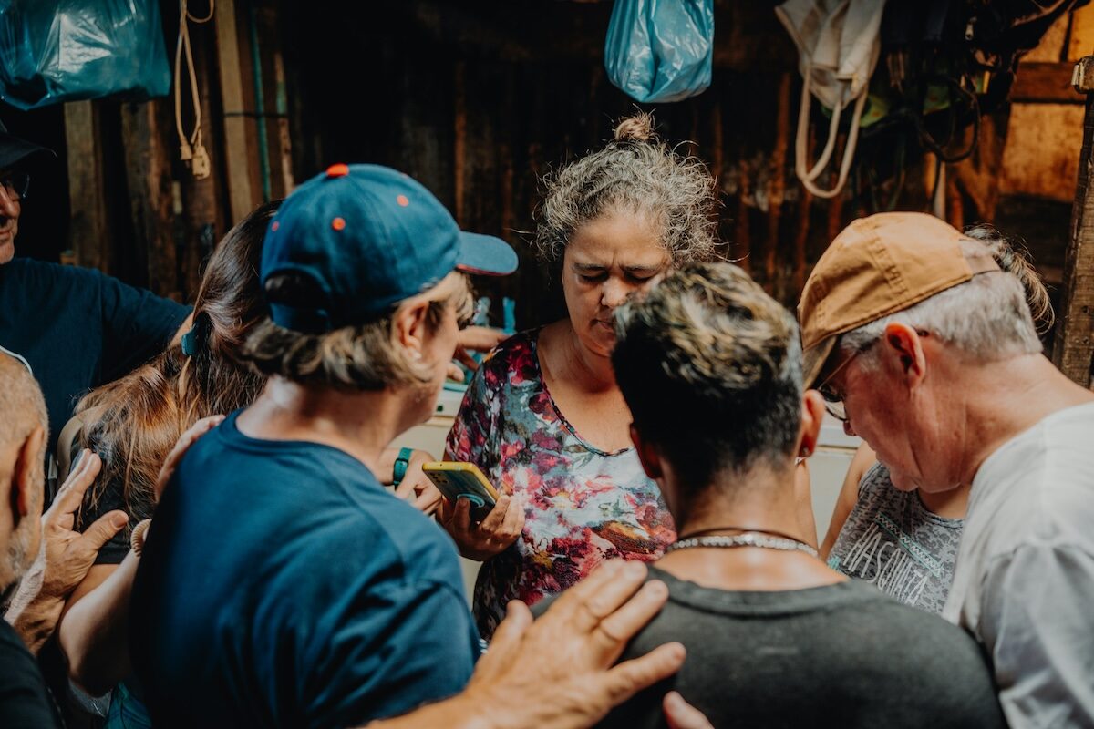 A group of people gather closely in a circle indoors, some with hands on each others shoulders, appearing to participate in a shared activity or moment.