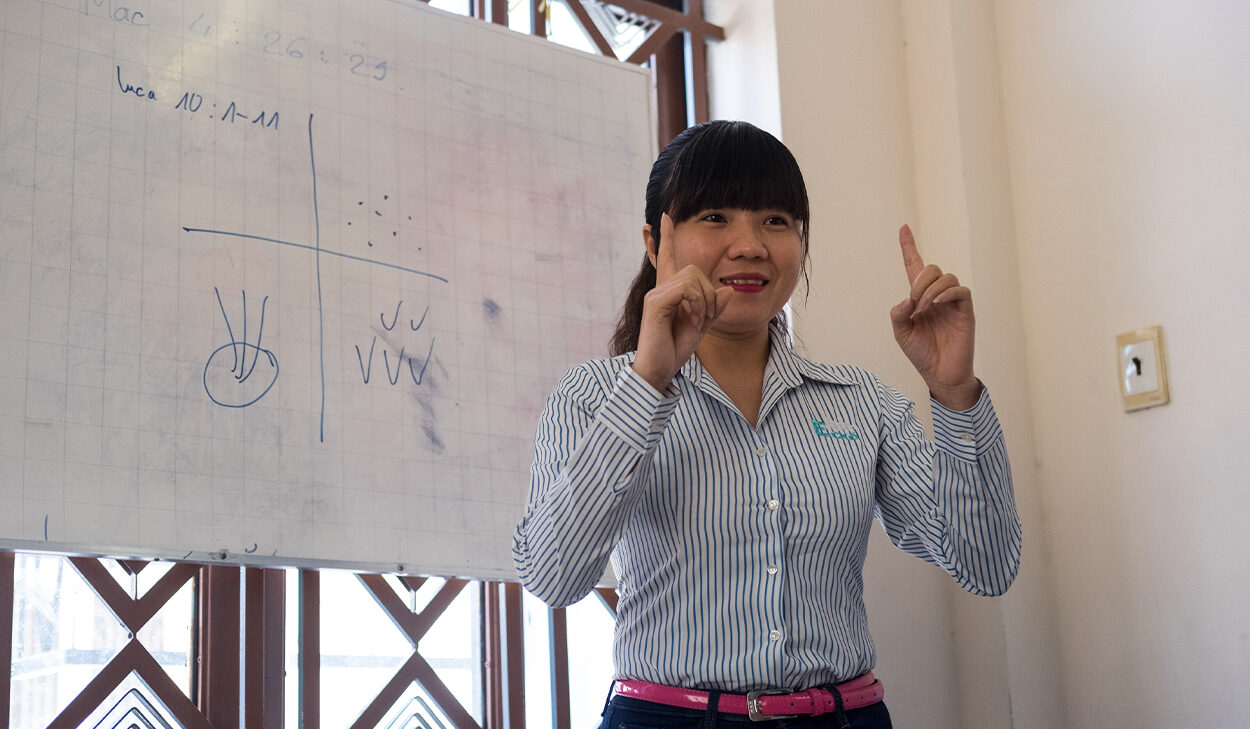 A woman in a striped shirt stands in front of a whiteboard, teaching the Four Fields.