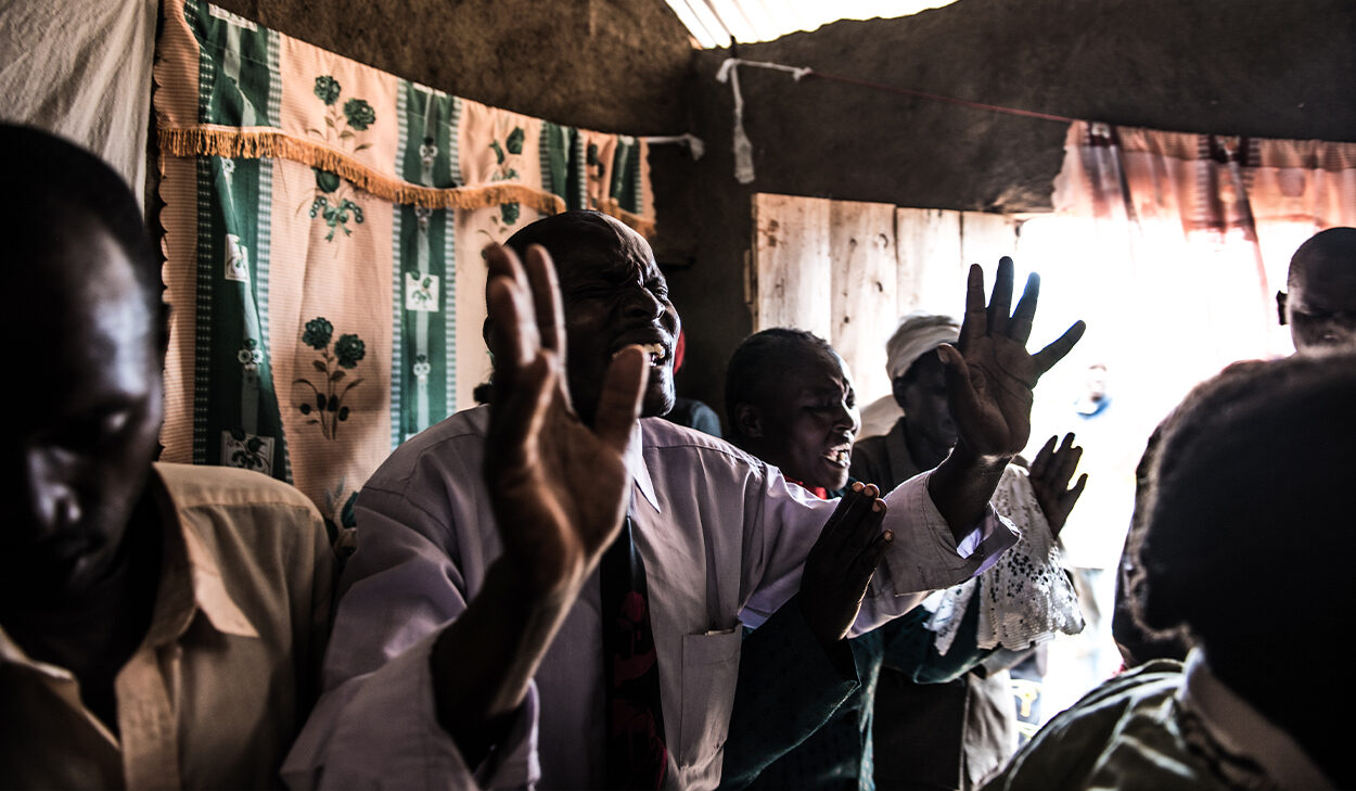A man wearing a white shirt and tie standing in an African church, singing and lifting his hands in praise.