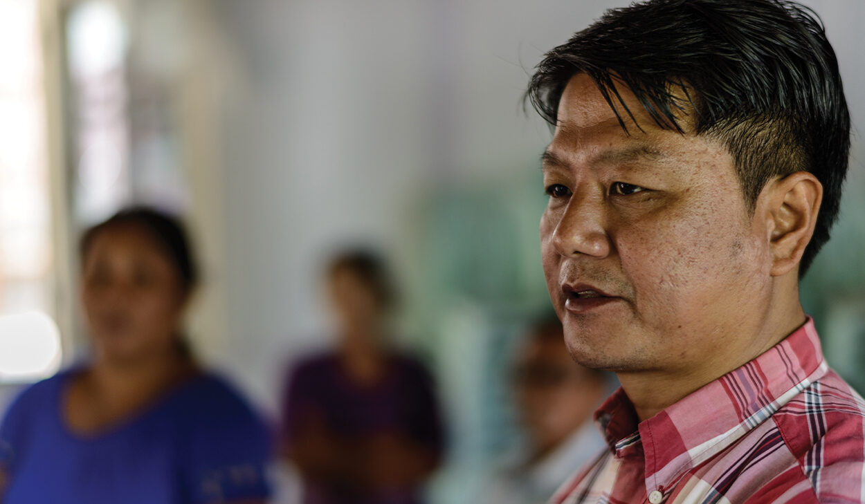 A man in a checkered shirt stands in a room. Other people are blurred in the background.