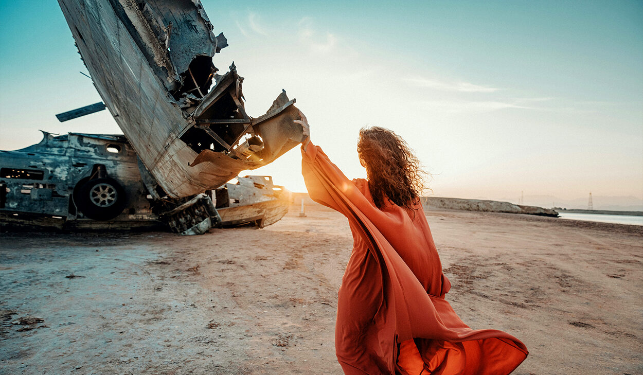 A woman in a rust-colored dress stands with her hand touching wreckage on a beach.