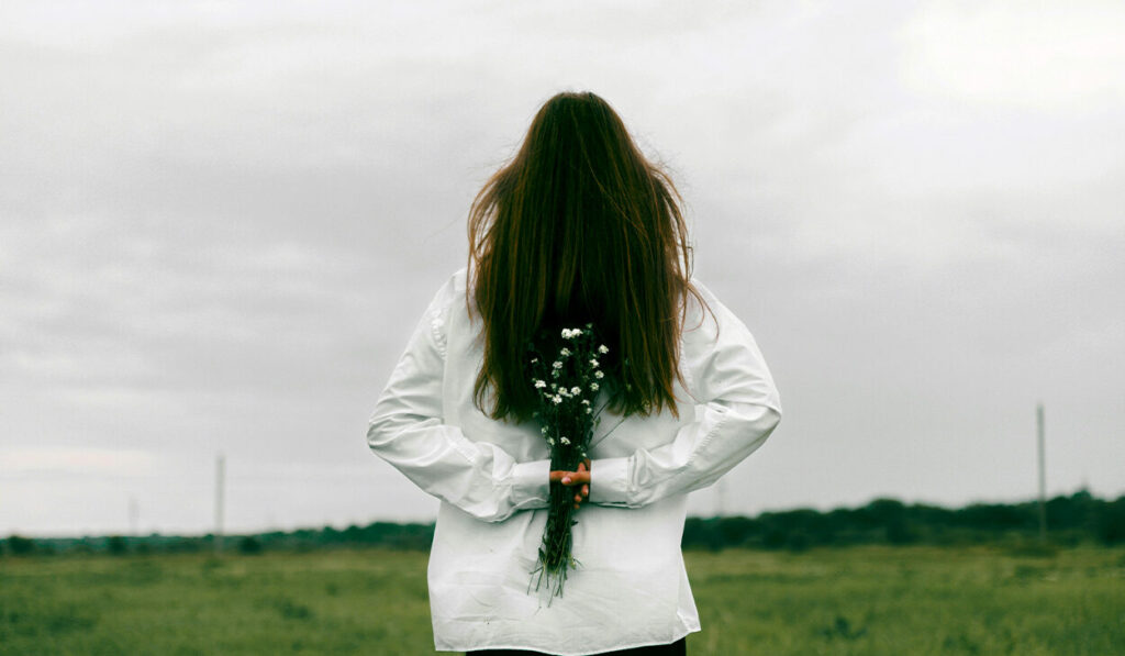 A woman with long, brown hair in a white sweater has her back to the camera, facing a green field. She is holding small, white flowers behind her back.