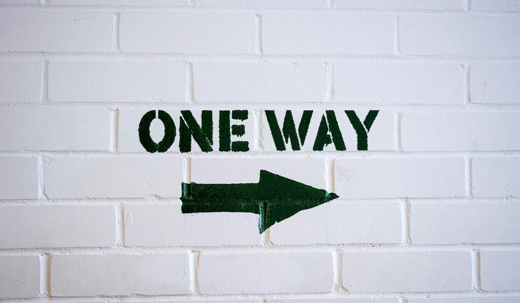 A one way arrow is painted in black on a white brick wall.