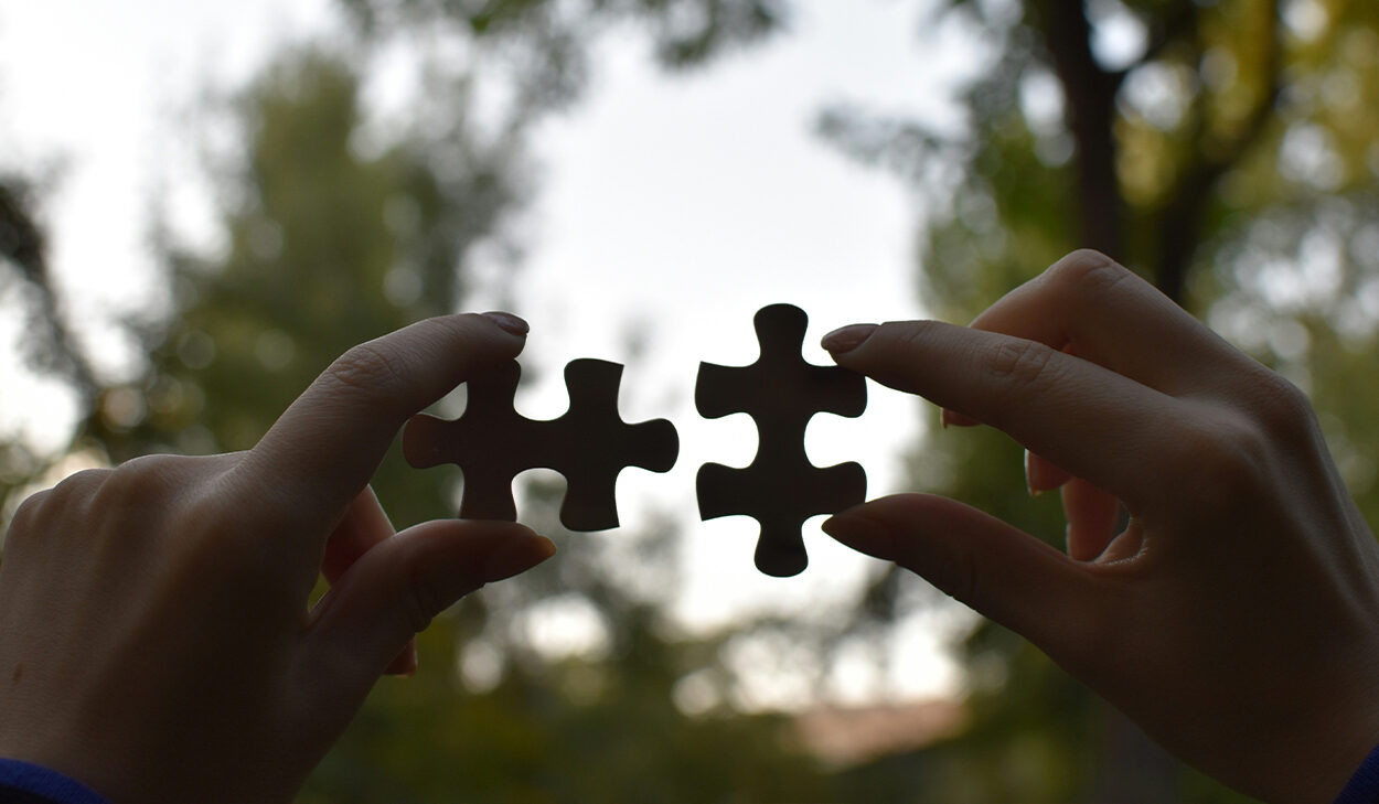 A woman holds up two puzzle pieces that fit together while standing in a wooded area.