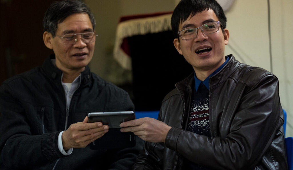 Two men in black jackets and glasses smile while one looks at a phone.