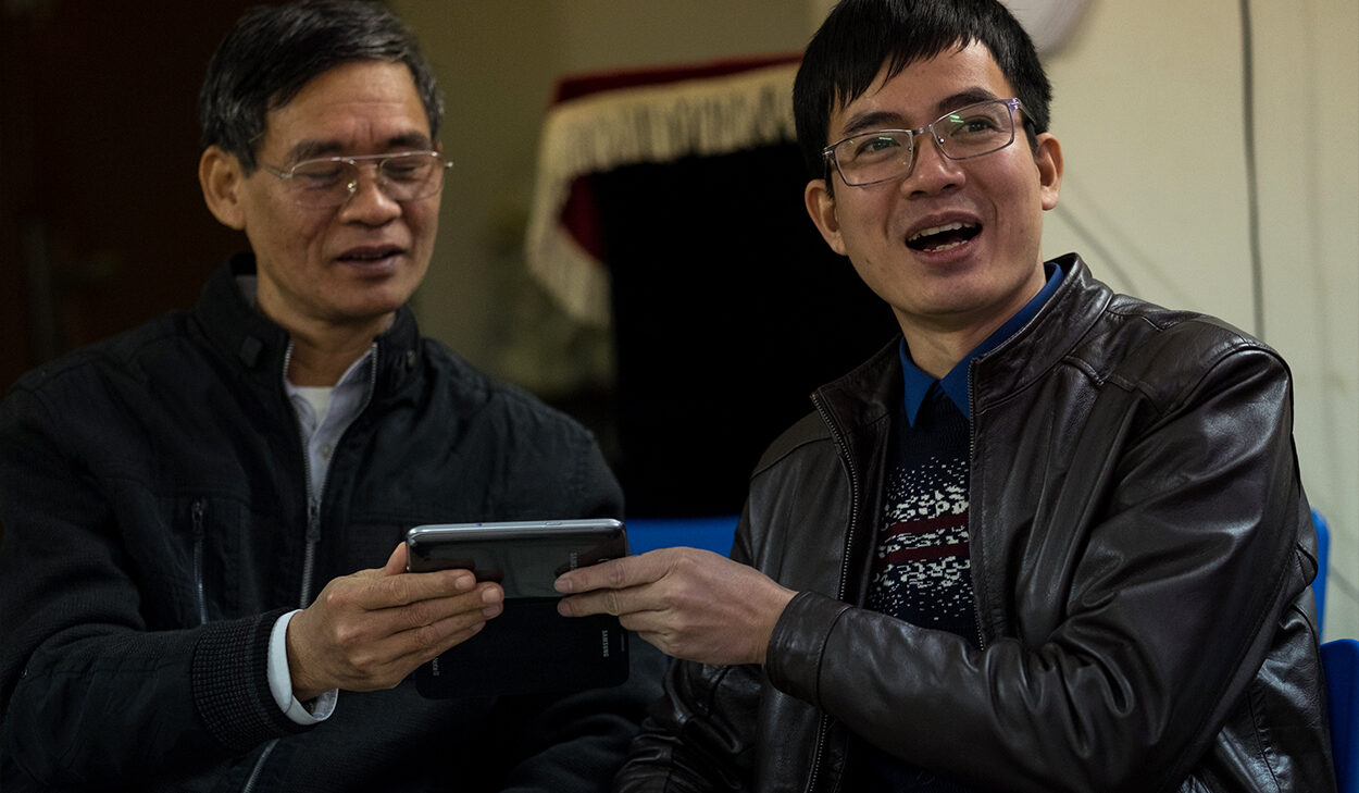 Two men in black jackets and glasses smile while one looks at a phone.