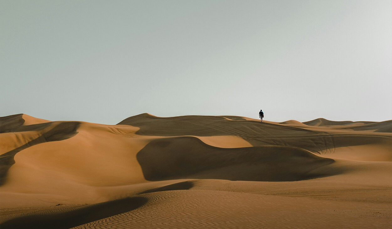 A person walks along sand dunes. The sky is clear.