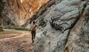 A woman in hiking gear walks through a canyon, brushing her hand against the rock as she walks.