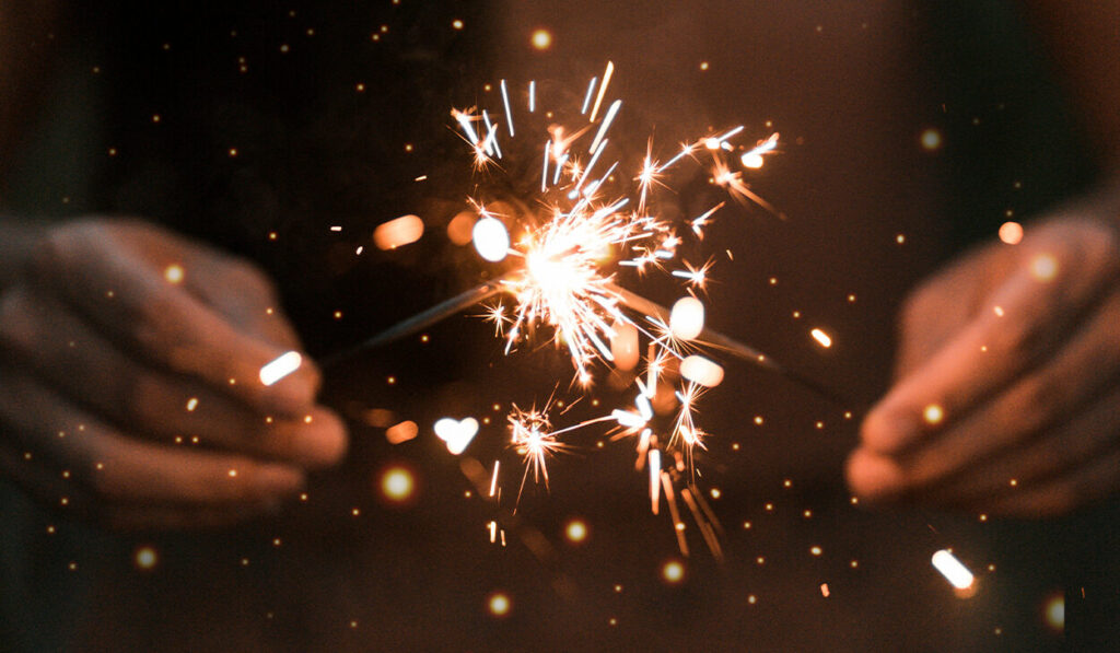 A person holds two lit sparklers in front of him.