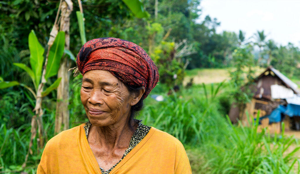 An elderly woman with a red scarf on her head and a yellow shirt stands in front of a lush, green background with a hut.