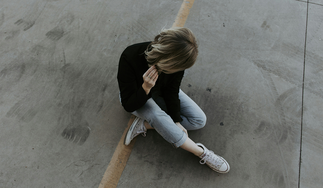 A woman in a black shirt sits in the street with her head down. Her face is hidden from the camera.