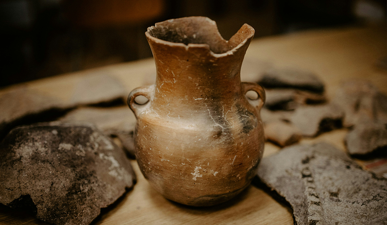 A cracked jar sits on a table surrounded by other pieces of broken pottery.