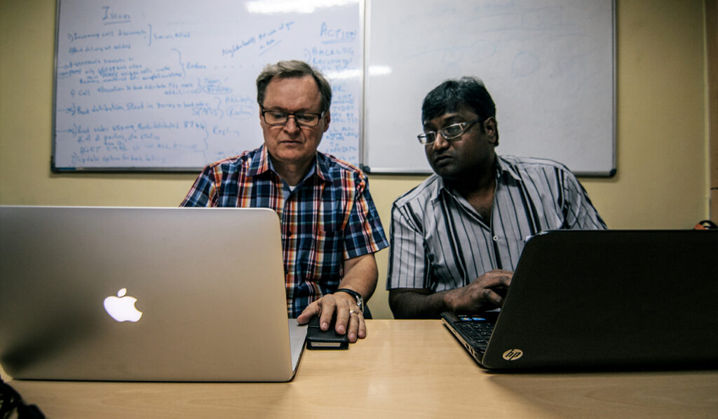 Two men sit side by side, looking at data on laptops.