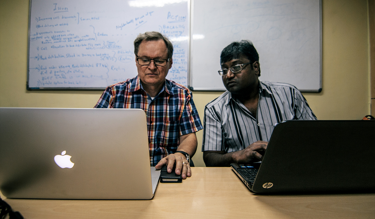 Two men sit side by side, looking at data on laptops.