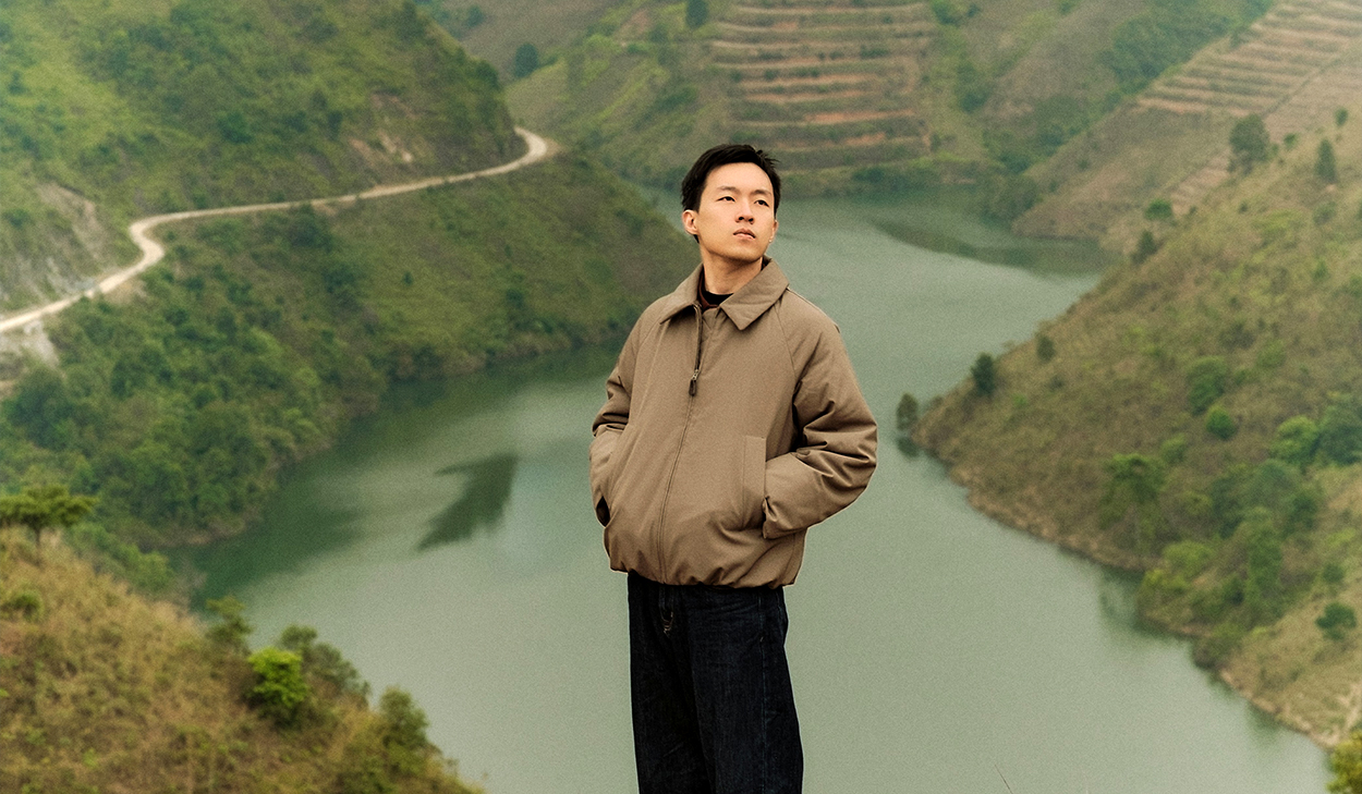 A man in a tan jacket and blue jeans stands in front of a lake in Southeast Asia, looking up at the sky.
