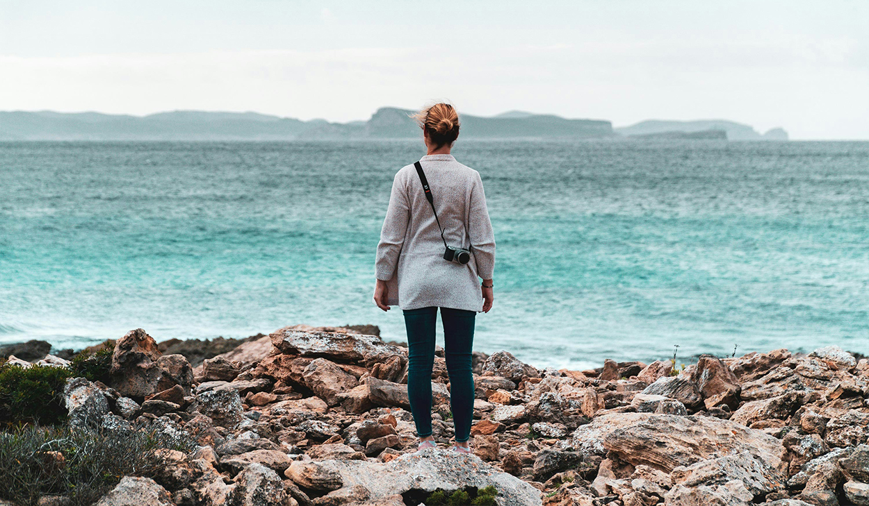 A woman in a sweater stands on a rocky beach overlooking the ocean. She has a camera strapped around her shoulder.