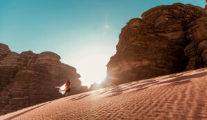 east-west-Arise 27 A woman walks up a sand dune to a rock formation as the sun peeks through the rocks.