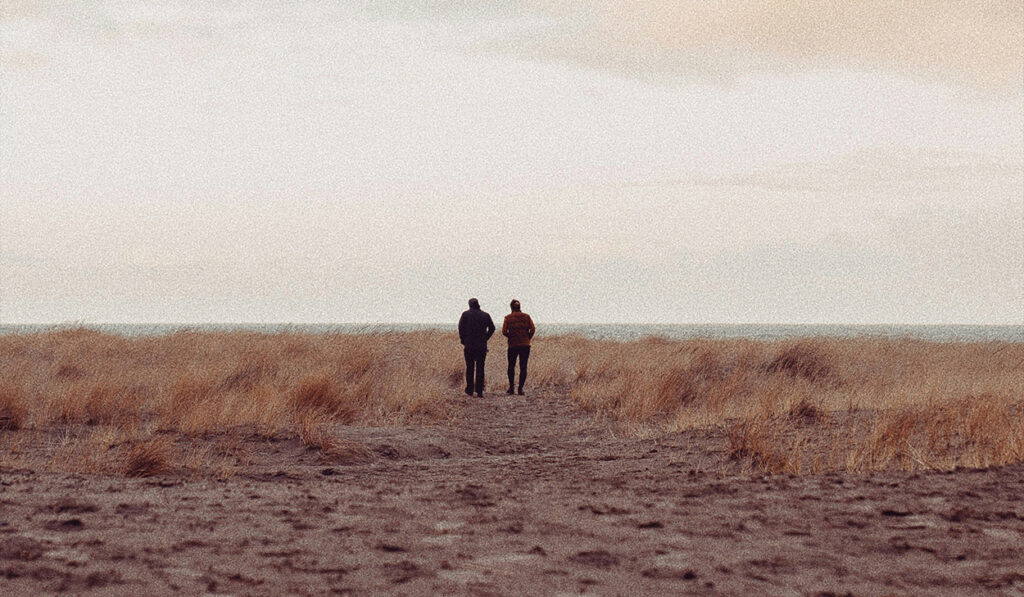 Two people walk toward the ocean on a grassy beach.
