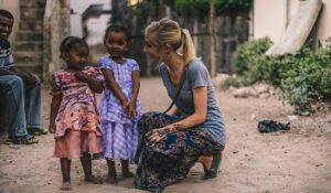 east-west-Arise 27 A woman in a skirt kneels next to two African girls on a dirt road.