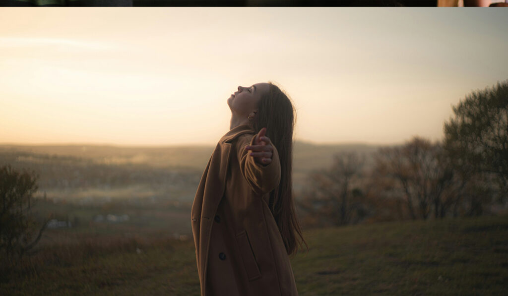 A woman in a coat stands with her arms stretched out and her face turned up toward the sky.