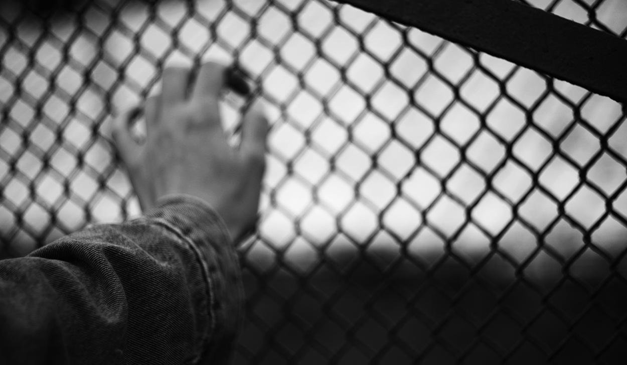 A black and white photo of a hand on a chain-link fence.