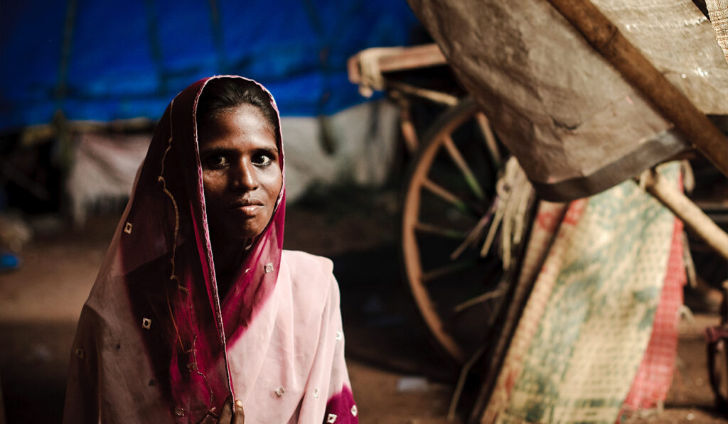 A South Asian woman in a rural village looks at the camera.
